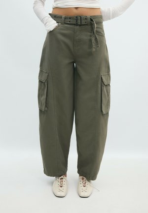 Femme portant un pantalon cargo taille haute vert olive à jambes larges avec ceinture et chaussures blanches à lacets, debout les mains dans les poches.