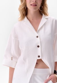 Witte overhemd met korte mouwen en een kraag, voorzien van bruine knopen en een gemêleerde stof. Draagt over hoge witte broeken.