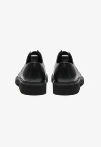 Zapatos de cuero negro con un acabado liso, punta redondeada y suela de goma texturizada en contraste. Pequeñas lengüetas en el talón para facilitar el calzado.