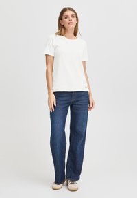 T-shirt bianco a maniche corte con colletto rotondo, abbinato a jeans a gamba larga blu scuro. Il tessuto sembra avere una certa texture, con dettagli discreti del marchio.