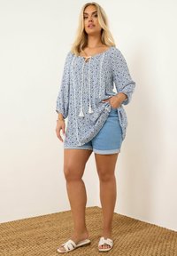 Kukkaroseläpitsi sinivalkoisessa kukkakuosisessa tunikassa, jossa on 3/4 hihat ja solmittava yksityiskohta. Yhdistetty vaaleisiin farkkushortseihin ja valkoisiin sandaaleihin. Teksturoitu tausta.