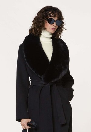 Frau mit lockigem Haar, die einen schwarzen Mantel mit Pelzkragen trägt, einen weißen Rollkragenpullover, schwarze Sonnenbrille und einen schwarzen Gürtel hält, die eine Hand in der Tasche hat.