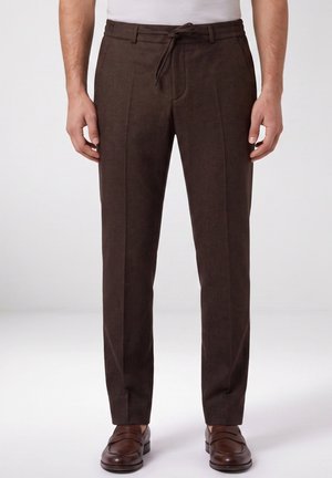 Man draagt bruine, op maat gemaakte broek met scherpe vouw vooraan, taillekoorden, wit overhemd en bruine leren loafers, staand tegen een effen achtergrond.
