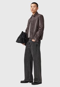 Homme portant une veste en cuir marron foncé, une chemise grise, un jeans noir ample, des chaussures noires, tenant un sac noir devant un arrière-plan clair.