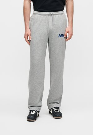 Homme portant un pantalon de survêtement Nike gris clair avec taille à cordon et des baskets noires à lacets blancs, debout devant un fond uni.
