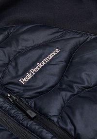 Zwarte gewatteerde jas met een gequiltde textuur, Peak Performance-logo in lichtroze en een ritszakdetail.