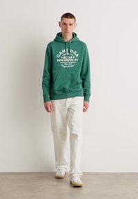 Hanorac verde din bumbac cu text alb, buzunar frontal și șnur ajustabil. Asortat cu pantaloni albi și adidași ușori.