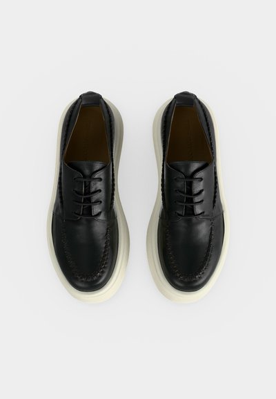 Μαύρα δερμάτινα παπούτσια με κορδόνια και χοντρές λευκές λαστιχένιες σόλες, με ραμμένες λεπτομέρειες στην περιοχή των δακτύλων, απεικονίζονται από πάνω σε ανοιχτό φόντο.