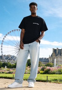 Jeune homme portant une chemise noire et un jean clair se tient dans un parc avec une grande roue et des bâtiments historiques derrière lui sous un ciel bleu.
