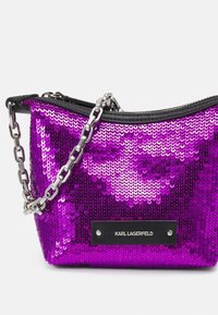 Fioletowa torebka z cekinami o zaokrąglonym kształcie, czarnym zamkiem błyskawicznym i srebrnym łańcuszkiem; z przodu znajduje się czarna metka z logo marki.