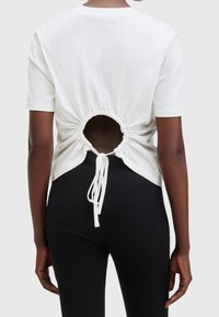 Zadní pohled na osobu v bílém krátkém rukávu s kruhovým výřezem a vázáním na dolní části zad, v kombinaci s černými ribbed kalhotami.