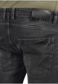 Jean en denim noir avec un patch en cuir à la taille, comprenant deux poches arrière et un détail de couture visible. La texture semble usée.