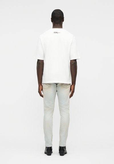Άνδρας που στέκεται πλάτη φορώντας λευκό κοντομάνικο μπλουζάκι με το κείμενο «ICONique» και ανοιχτόχρωμα τζιν με μαύρα παπούτσια.