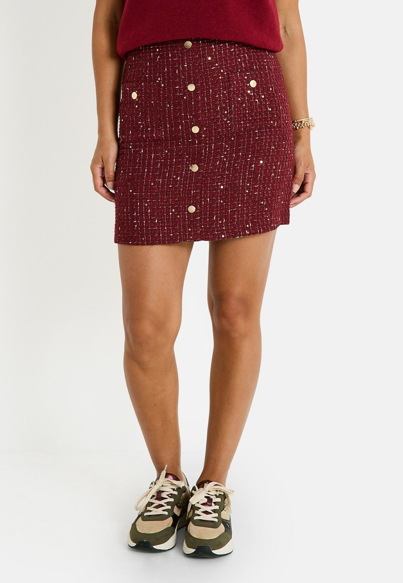 Falda roja texturizada con acentos metálicos, botones frontales y una forma ajustada, combinada con zapatillas beige y verdes.