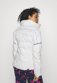 Weiße gepolsterte Jacke mit hohem Kragen, verziert mit blauen Paspeln. Getragen mit Hosen im Blumenmuster, die eine glatte, glänzende Textur aufweisen.
