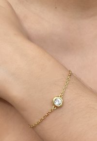 Bracciale d'oro con una catena delicata, presenta una pietra centrale trasparente incastonata in una montatura circolare. Texture liscia, design minimalista.