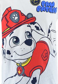 Witte t-shirt met een cartoon hond in rode brandweeruitrusting. Het ontwerp bevat zwarte vlekken en een pootlogo op het shirt.