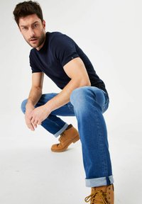 Camiseta de manga corta azul marino, jeans azul claros y botas marrones con cordones amarillos. Modelo agachado con las manos reposando sobre las rodillas, fondo neutro.