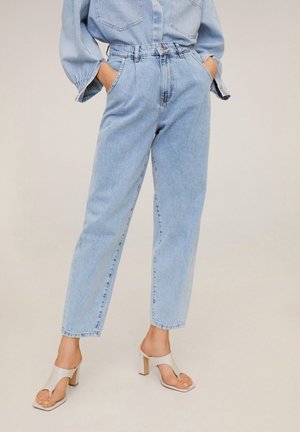 Persona con jeans de tiro alto azul claro y camisa de mezclilla a juego, con las manos en los bolsillos, combinados con sandalias beige de tacón ancho y punta abierta.