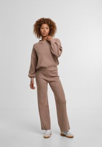 Pull en maille gaufrée marron clair avec manches ballon, associé à un pantalon taille haute à jambes larges assorti. Des baskets blanches complètent la tenue.