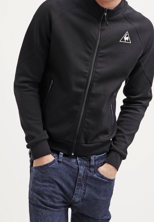 Person trägt eine schwarze Jacke mit Reißverschluss und einem weißen dreieckigen Logo auf der Brust sowie blaue Jeans, Hände in den Taschen, steht vor einem einfarbigen Hintergrund.