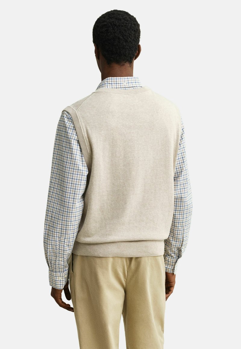 Homme portant un gilet en tricot beige par-dessus une chemise à carreaux bleue et blanche et un pantalon beige, vu de dos sur un fond uni.