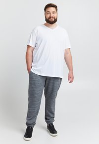 Witte v-hals t-shirt en grijze joggingbroek, textuurrijke stof. Zwarte sneakers maken de casual outfit compleet. Model staat met één hand in de zak.