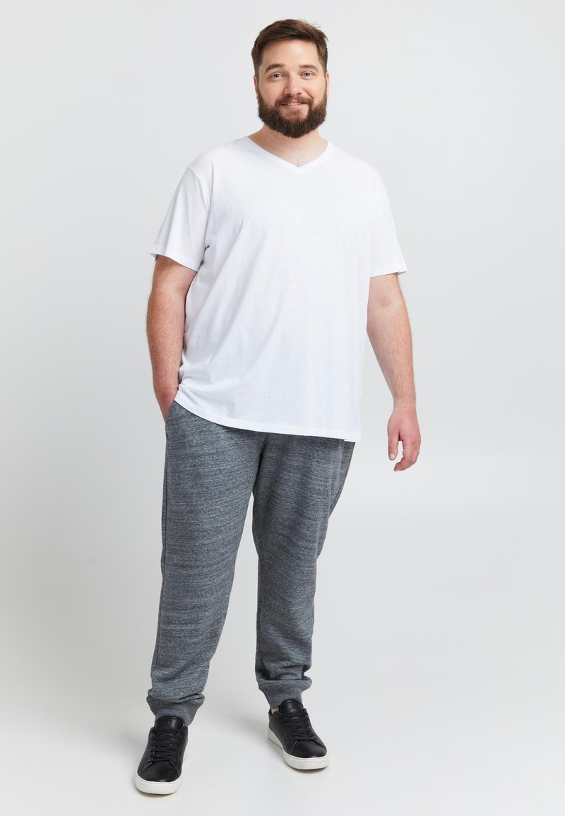 Witte v-hals t-shirt en grijze joggingbroek, textuurrijke stof. Zwarte sneakers maken de casual outfit compleet. Model staat met één hand in de zak.