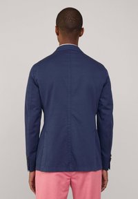 Granatowa kurtka o dopasowanym kroju, z pojedynczym rozcięciem i guziczkami na mankietach. Gładki materiał z minimalistycznym wzornictwem, zestawiona z różowymi spodniami.