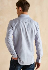 Koszula z guzikami w niebiesko-białe pionowe paski, wykonana z bawełny. Posiada ostry kołnierzyk i mankiety zapinane na guziki. Logo marki na plecach.