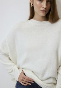 White knit sweater with a round neck and ribbed cuffs, featuring a relaxed fit and soft texture, worn with dark jeans.