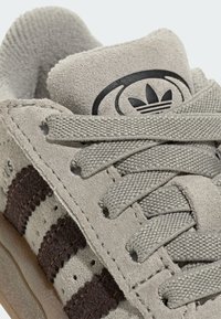 Close-up of beige suede sneaker with brown stripes, detailed laces, and Adidas trefoil logo on the padded tongue.