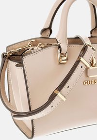Sac à main en synthétique beige avec quincaillerie dorée, anses doubles et une bandoulière amovible. Présente des accents de couture et le nom de la marque affiché de manière proéminente.