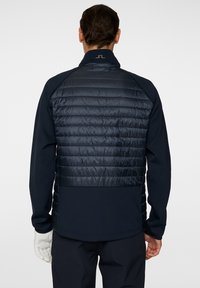 En person står med ryggen mot kameran, iklädd en mörk marinblå jacka med ett quiltat mönster och en logotyp på kragen.