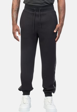 Pantaloni jogger neri con vita con coulisse, polsini a coste e texture liscia, abbinati a sneakers bianche.