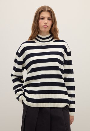 Giovane donna con lunghi capelli ondulati che indossa un maglione a collo alto a righe orizzontali bianche e nere e pantaloni neri, in piedi contro una parete liscia.