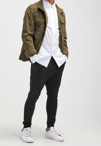 Olive green zip-up jacket over a white button-up shirt, paired with black tapered joggers and white sneakers.