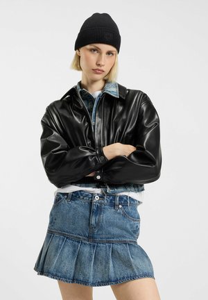 Giacca nera in ecopelle con colletto strutturato, indossata sopra una camicia di jeans azzurro chiaro e una gonna di jeans a pieghe. Berretto nero in maglia.