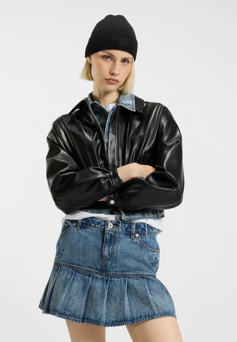 Czarna kurtka z eko-skóry z kanciastym kołnierzem, noszona na jasnoniebieskiej koszuli jeansowej i plisowanej spódnicy jeansowej. Czarna dziana czapka.