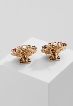 Goudkleurige clip-on oorbellen met meerkleurige vierkante edelstenen gerangschikt in een cirkelvormig patroon, tentoongesteld op een witte kubus.