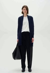 Mørkeblå lang cardigan laget av mykt stoff, åpen front, kombinert med en hvit skjorte og løse svarte bukser. Modellen holder en stor svart veske.