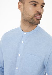 Camisa de lino de color azul claro con escote redondo, cierre de botones y un bolsillo en el pecho. Presenta una textura suave y un sutil patrón jaspeado.