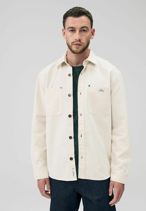 Hombre vistiendo una camisa beige desabotonada sobre una camiseta verde oscura y jeans oscuros, de pie frente a un fondo claro y liso.
