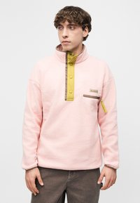 Νεαρός άνδρας φορώντας ροζ φλις πουλόβερ με κίτρινη κλειστή λωρίδα κουμπιών και τσέπη με φερμουάρ στο στήθος, στέκεται μπροστά σε απλό λευκό φόντο.