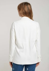 Blazer blanc à coupe classique, en tissu doux, avec des épaules structurées et des accents de boutons aux poignets, associé à un jean bleu.