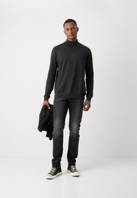 Mørkegrå turtleneck sweater, slim-fit sorte jeans, sorte sneakers med hvide såler, holder en sort læderjakke. Glat tekstur, afslappet design.
