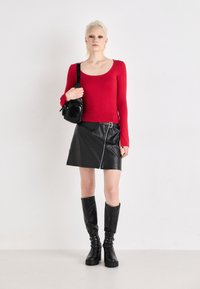 Rotes Langarmoberteil, schwarze Kunstleder-Rock mit Reißverschluss, knielange schwarze Stiefel mit Nieten und eine kleine schwarze Umhängetasche. Einfarbiger Hintergrund.