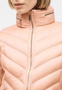 Nærbillede af en person iført en lyserød quiltet jakke med høj krave og guldfarvede lynlåse, delvist lynet op.