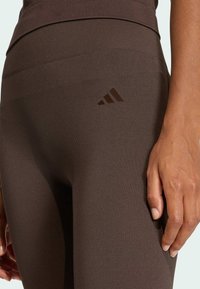 Hnědé atletické legíny s jemně vroubkovanou strukturou a vysokým pasem. Na boku se nachází tmavě hnědé logo Adidas. Detailní záběr ukazuje ruku odloženou na látce.