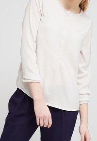 Camicia a maniche lunghe color crema con scollatura rotonda, taschino sul petto e orlo leggermente curvo. Tessuto morbido e vestibilità comoda con maniche arrotolate.
